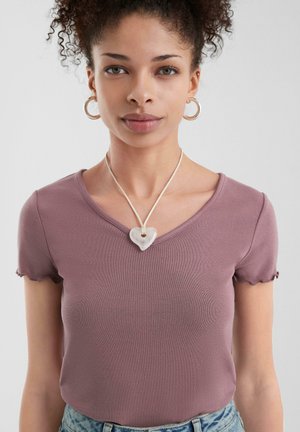 Camiseta de rayas moradas con mangas cortas y bordes ondulados, combinada con un colgante de corazón en un cordón de cuero. La modelo lleva aretes tipo aro.