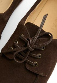 Scarpa in suede marrone con dettagli di allacciatura, caratterizzata da occhielli in metallo e un bordo cucito. L'interno presenta una fodera beige e un design minimalista.
