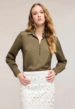 Jeune femme aux cheveux blonds longs et ondulés, portant un chemisier manches longues vert olive et une jupe blanche à motifs, les mains posées sur les hanches.