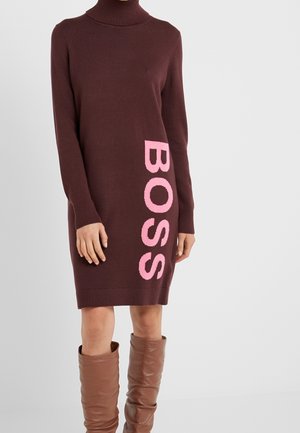 Osoba nosząca brązową sukienkę z golfem z napisem „BOSS” dużymi różowymi literami oraz luźne brązowe skórzane kozaki do połowy łydki.