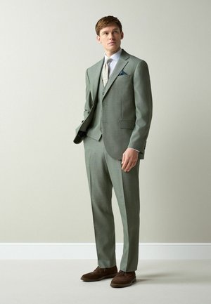 Muž oblečený v svetlosivom trojdielnom obleku, bielej košeli, svetlosivej kravate a hnedej obuvi, stojaci pred jednoduchou svetlou stenou.