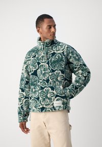 Maglione morbido in teal e crema con un motivo floreale, colletto alto e chiusura con zip. Presenta tasche laterali e etichetta del marchio sull'orlo.