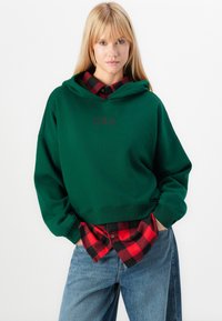 Sweat à capuche vert avec une poche avant, arborant un logo "GAP" sombre. Associé à une chemise à carreaux noire et rouge portée en dessous. Jeans bleus.