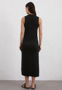 Schwarzes, ärmelloses gestricktes Maxikleid mit hohem Kragen und seitlichen Schlitzen. Glatte Textur, figurbetonte Silhouette, die bis zur Mitte der Wade reicht. Getragen mit flachen Sandalen.