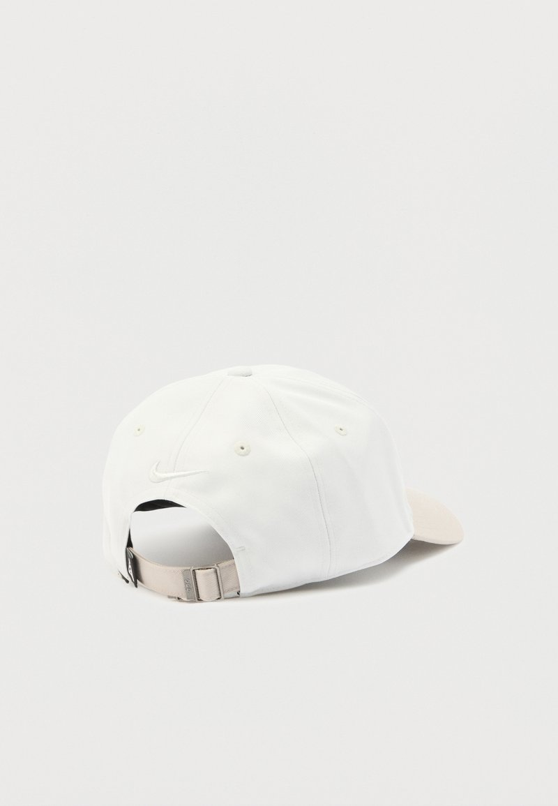 Weißes Baseballcap mit verstellbarem beigem Riemen und kleinem Nike-Swoosh-Logo auf der Rückseite, von hinten auf hellem Hintergrund gezeigt.