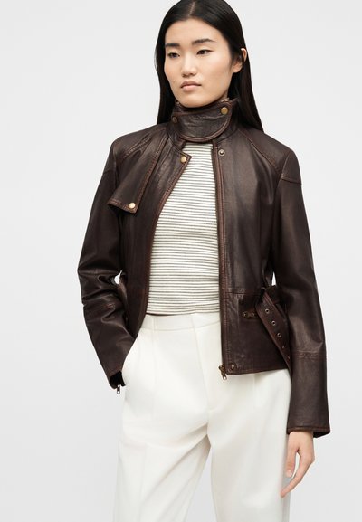 Γυναίκα που φοράει σκούρο καφέ δερμάτινο μπουφάν πάνω από ένα λευκό ριγέ πουκάμισο και λευκό παντελόνι ψηλόμεσο, στέκεται με το ένα χέρι στην τσέπη.