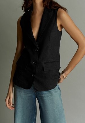 Femme portant un blazer noir sans manches boutonné et un jean bleu clair sur un fond gris uni.