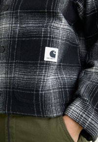 Crna i siva karirana flanel košulja s bijelom etiketom koja sadrži crni logo. Vidljivi su dugmići i teksturirana tkanina, uz detalj džepa.