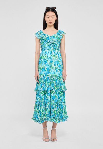 Robe maxi sans manches à fleurs dans des nuances de bleu et de vert, avec des manches volantées et des couches au niveau de l'ourlet. Sandales à talons transparentes et à lanières.