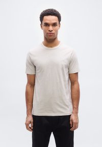 Ανοιχτό μπεζ κοντομάνικο μπλουζάκι με στρογγυλή λαιμόκοψη και διακριτικό λογότυπο στο στήθος, κατασκευασμένο από μαλακό, λείο ύφασμα. Συνδυάστε το με σκούρο παντελόνι.