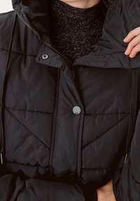 Svart quiltad pufferjacka med huva, med tryckknappsstängning och dragsko. Under är en texturerad mörk stickad tröja synlig.