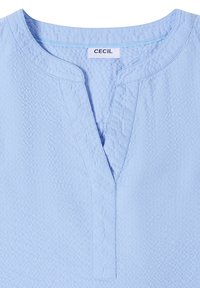 Haut en tissu bleu clair texturé avec un col rond fendu et une étiquette de marque "CECIL" visible à l'intérieur du col.