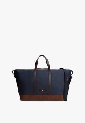 Granatowa torba typu duffel z brązową skórzaną podstawą i uchwytami, zapinana na zamek u góry, z regulowanym paskiem na ramię, z małym prostokątnym logo.