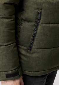 Grøn quiltet pufferjakke med en sidelomme med lynlås, tekstureret stof og Velcro-manchetdetaljer. Nærbillede af ærme- og lomme detaljer.