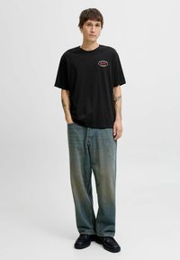 Zwarte katoenen t-shirt met een kleine rode en witte graphic op de borst, gecombineerd met baggy vervaagde blauwe jeans en zwarte schoenen.