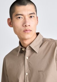 Camisa abotonada de color marrón claro, fabricada en una tela suave, que presenta un cuello clásico y un logo de la marca bordado en el pecho. Textura suave.