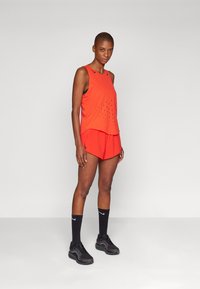 Rotes sportliches Tanktop mit perforiertem Design, kombiniert mit roten Shorts. Das Model trägt schwarze Socken und schwarze Laufschuhe. Einfacher Hintergrund.