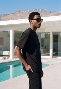 Uomo con t-shirt nera e occhiali da sole in piedi vicino a una piscina con una casa bianca moderna e montagne sullo sfondo.