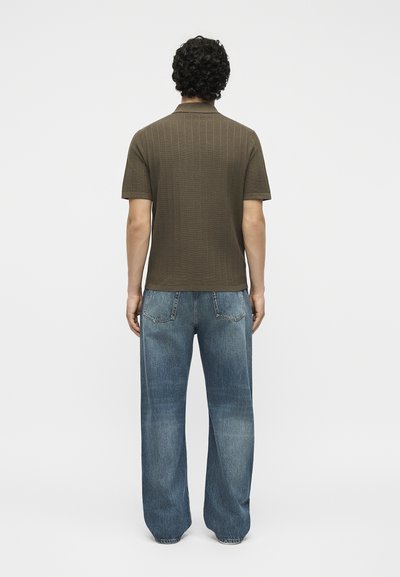 Άνδρας με την πλάτη, φοράει καφέ πλεκτή πόλο μπλούζα με κοντό μανίκι και άνετο μπλε τζιν παντελόνι, σε απλό λευκό φόντο.