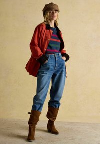 Červená nepremokavá bunda, pruhované modré a zelené tričko, modré rifle s vysokým pásom a hnedé suède čižmy po kolená. Hladká hnedá čiapka.