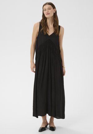 Femme portant une robe maxi noire sans manches avec un col en V et des détails en franges, associée à des chaussures à talons noires sans lacets, debout sur un fond uni.