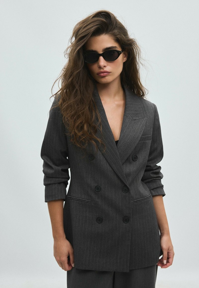 Femme aux longs cheveux ondulés porte des lunettes de soleil noires en forme d’œil de chat et un blazer croisé à fines rayures gris foncé avec les manches retroussées.
