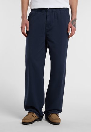 Man draagt navyblauwe losse broek, bruine schoenen en een wit overhemd, staand op een effen witte achtergrond.