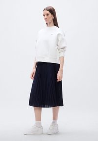 Sweat-shirt blanc à col rond et épaules tombantes, associé à une jupe plissée bleu marine et des baskets blanches. Design simple, texture lisse.