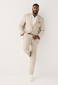 Homme barbu portant un costume beige, un polo blanc et des baskets blanches, debout sur une jambe devant un fond clair uni.