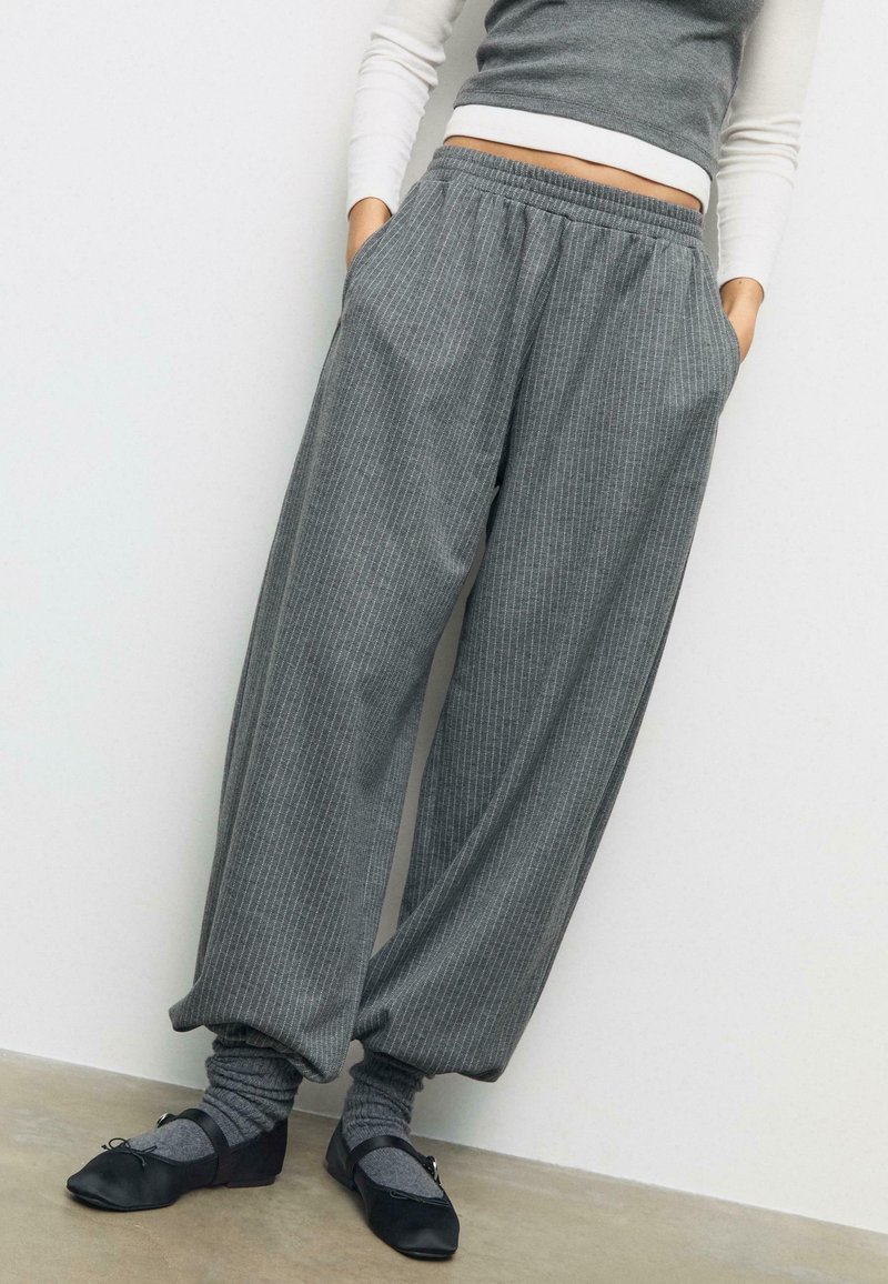 Svijetlosive, prugaste hlače opuštenog kroja s elastičnom trakom u struku, koje imaju bočne džepove, u kombinaciji s crnim balerinkama i sivim čarapama.