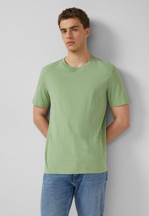 Junger Mann steht vor einer grauen Wand, trägt ein hellgrünes Kurzarm-T-Shirt und blaue Jeans, die Arme hinter dem Rücken verschränkt.