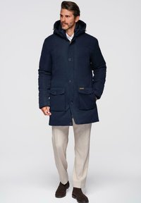 Marineblauwe waterdichte parka met een capuchon, knoopsluiting aan de voorkant, twee klepzakken en gouden hardwareaccenten. Kenmerkt zich door een relaxte pasvorm en een duurzame stoffen textuur.