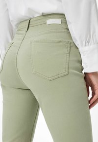 Hellgrüne Jeans aus elastischem Stoff, mit hoher Taille und Gesäßtaschen, die eine subtile strukturelle Oberfläche und kontrastierende Nähte aufweisen.