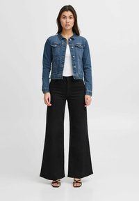 Chaqueta de mezclilla con un diseño ajustado, dos bolsillos en el pecho y un lavado claro. Combinada con pantalones negros de pierna ancha y sandalias negras con tiras.