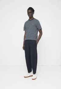 Άνδρας με κοτσίδες που φοράει μπλε ναυτικό και λευκό ριγέ T-shirt, μπλε παντελόνι και λευκά αθλητικά παπούτσια, στέκεται μπροστά σε απλό λευκό φόντο.