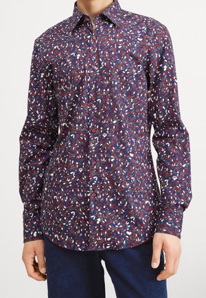 Mężczyzna w koszuli na długi rękaw zapinanej na guziki z niebieskim, czerwonym i białym abstrakcyjnym liściastym wzorem oraz w ciemnoniebieskich spodniach, stojący na białym tle.