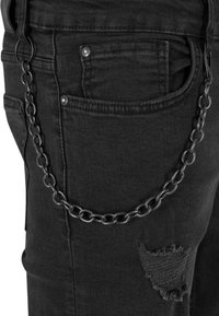 Jeans in denim nero con una catena prominente attaccata a un passante, presentano un dettaglio strappato nella zona della tasca destra.