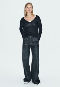 Schwarzer gestrickter Pullover mit weitem V-Ausschnitt und lockerem Sitz, kombiniert mit dunklen Jeans im Schlagstil, die kontrastierende weiße Nähte aufweisen.