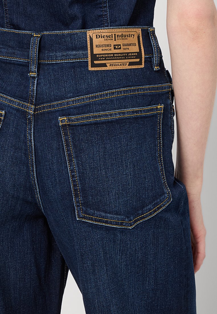 Dark blue denim overalls with a textured finish. Features include a tan leather label with brand details and contrasting yellow stitching.