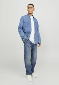 Hellblaue Jeanshemd mit zwei Brusttaschen, getragen über einem weißen T-Shirt. Lockere blaue Jeans und weiße Turnschuhe vervollständigen das Outfit.