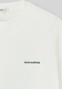 T-shirt blanc ample avec un col rond, présentant le mot "Untroubled" en texte vert foncé sur la poitrine. Tissu en coton doux.
