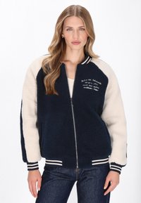 Marineblå og creme fleece bomberjakke med stribede manchetter, lynlås lukning og broderet tekst på forsiden. Blød tekstur, afslappet pasform.