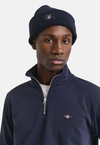 Marineblauwe zip-nekl pullover met een zachte textuur en het GANT-logo op de borst, gecombineerd met een bijpassende geribde breimuts.