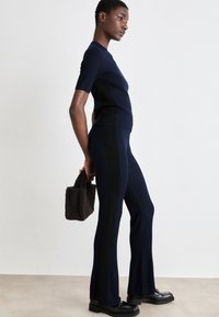 Top in maglia a coste blu navy e pantaloni svasati con pannelli laterali neri; indossato con una piccola borsa in pelliccia marrone scuro e scarpe nere.