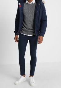 Man med marinblå jacka med pälsluva, grå tröja över vit skjorta, marinblå slim fit-byxor och vita sneakers står framför en neutral bakgrund.