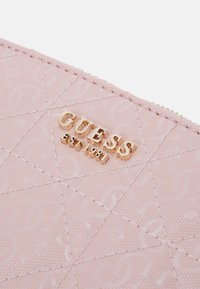 Różowy portfel z syntetycznej skóry z wypukłym wzorem diamentowym, złotym logo "GUESS" oraz subtelnymi detalami tekstury. Zamek błyskawiczny na górze.