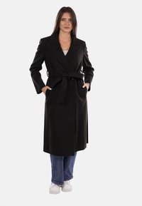 Donna che indossa un lungo cappotto nera con cintura, jeans blu e sneaker bianche, in piedi con le mani nelle tasche del cappotto su uno sfondo neutro.