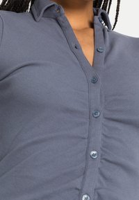 Grå randig knäppskjorta med krage och blåa knappar. Har texturerad tyg och samlingar på sidorna.