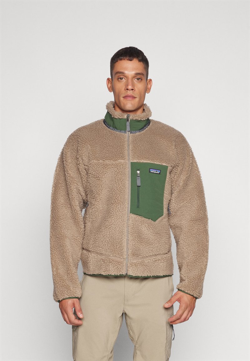 Tan fleece jacket with a green chest pocket, full zip, high collar, and textured surface. Features gray accents along the zipper.