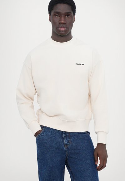 Sweatshirt beige clair avec un col rond et des poignets côtelés, doté d'un petit logo noir sur la poitrine. Assorti à un jean en denim bleu foncé.